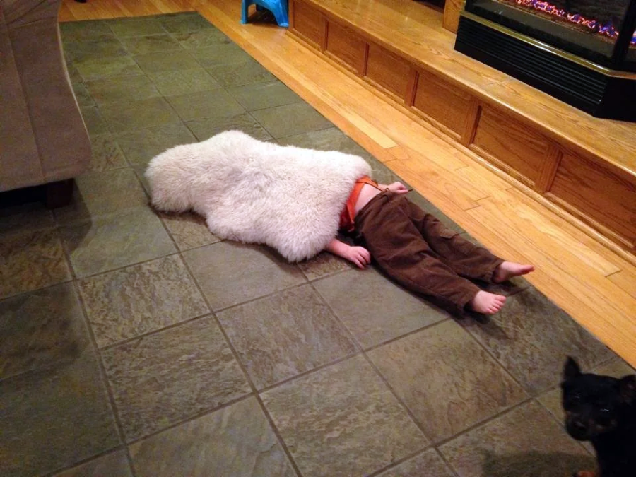 A child lies face down on a tiled floor with their head covered by a fluffy white rug, making it look like an animal. A small dog stands nearby, looking toward the camera.