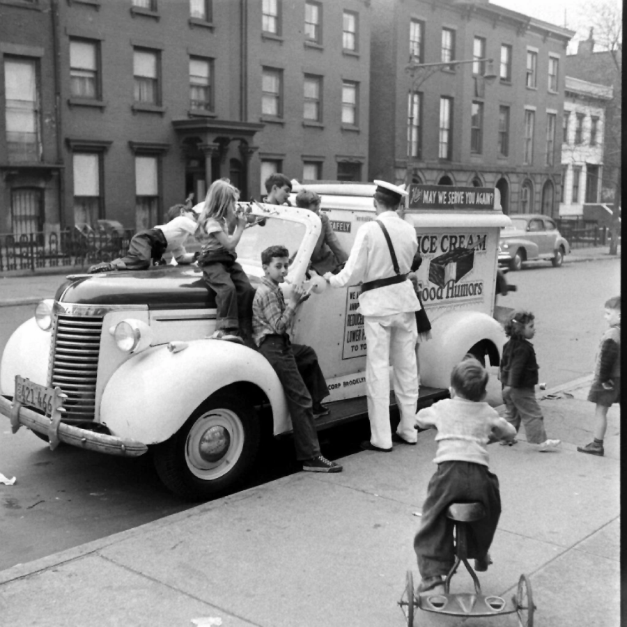 Children gather around a vintage ice cream truck with a uniformed vendor on a city sidewalk. Some kids climb onto the truck, while others wait nearby. An apartment building is in the background.