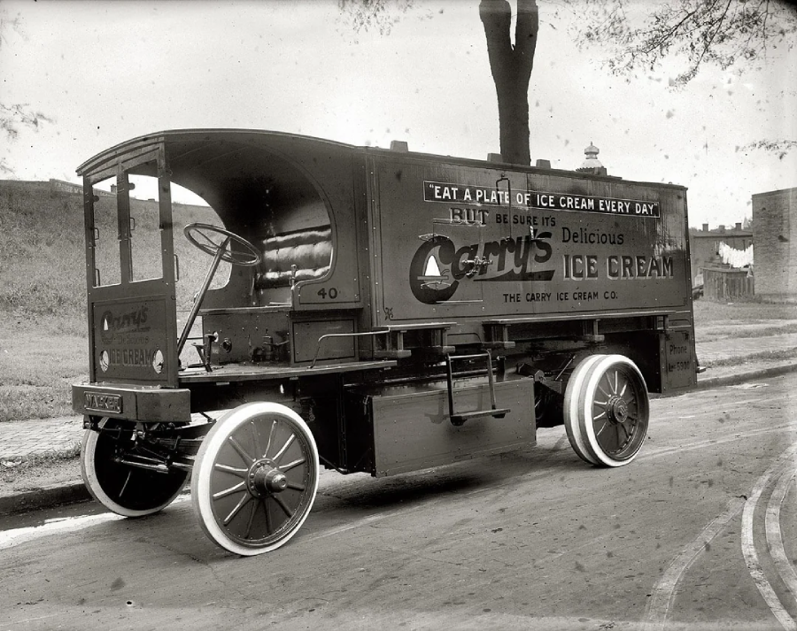 A vintage ice cream delivery truck with the sign “Cary’s Delicious Ice Cream” and a slogan about eating ice cream daily, parked on a street with trees and buildings in the background.