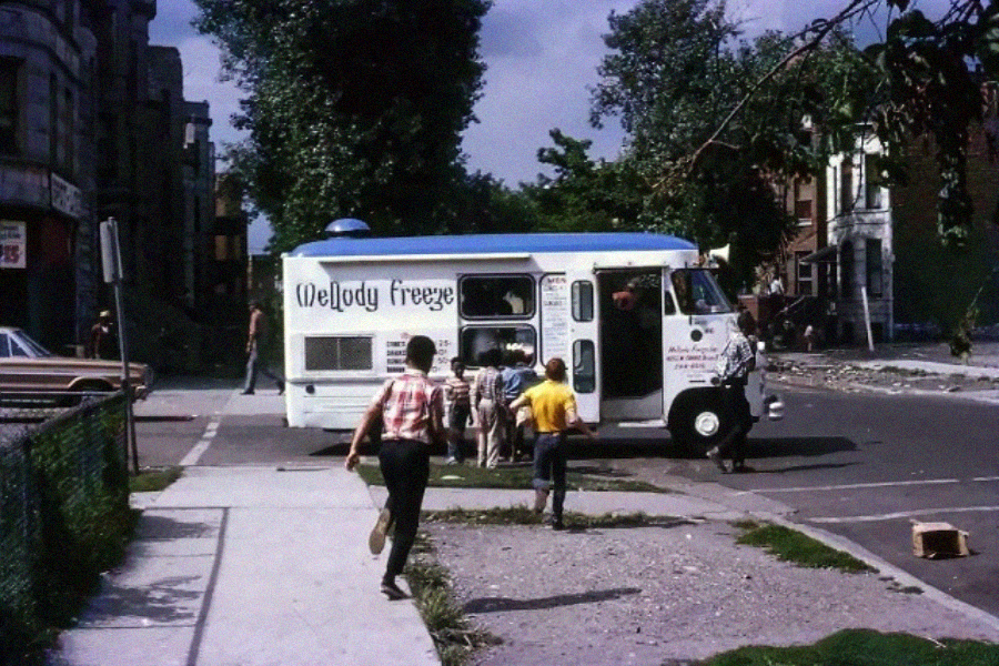 Children gather around a white and blue Melody Freeze ice cream truck parked on a city street, while one child runs toward it. Trees and buildings are visible in the background on a sunny day.