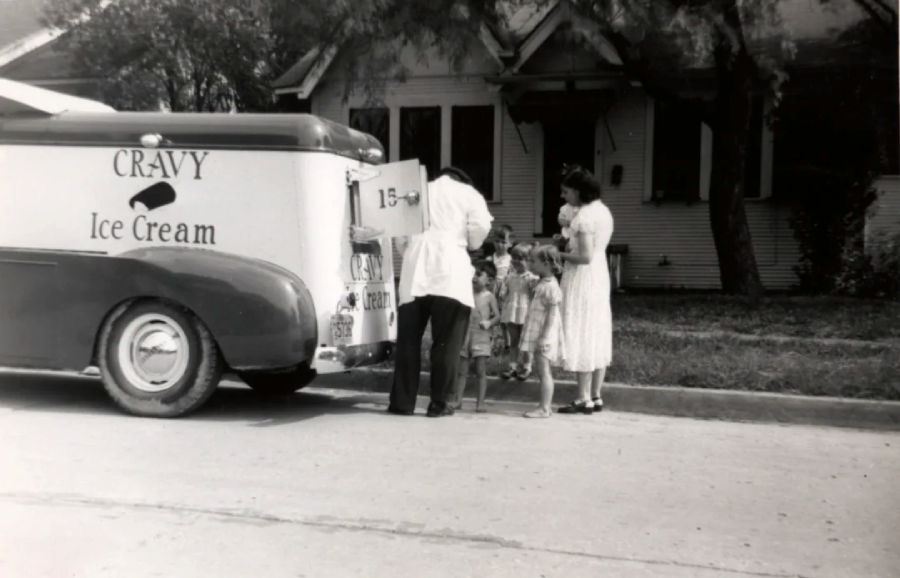 A black-and-white photo shows a vintage ice cream truck parked on a suburban street. A vendor serves ice cream to three children and a woman, all standing at the back of the truck near a house with trees in the background.