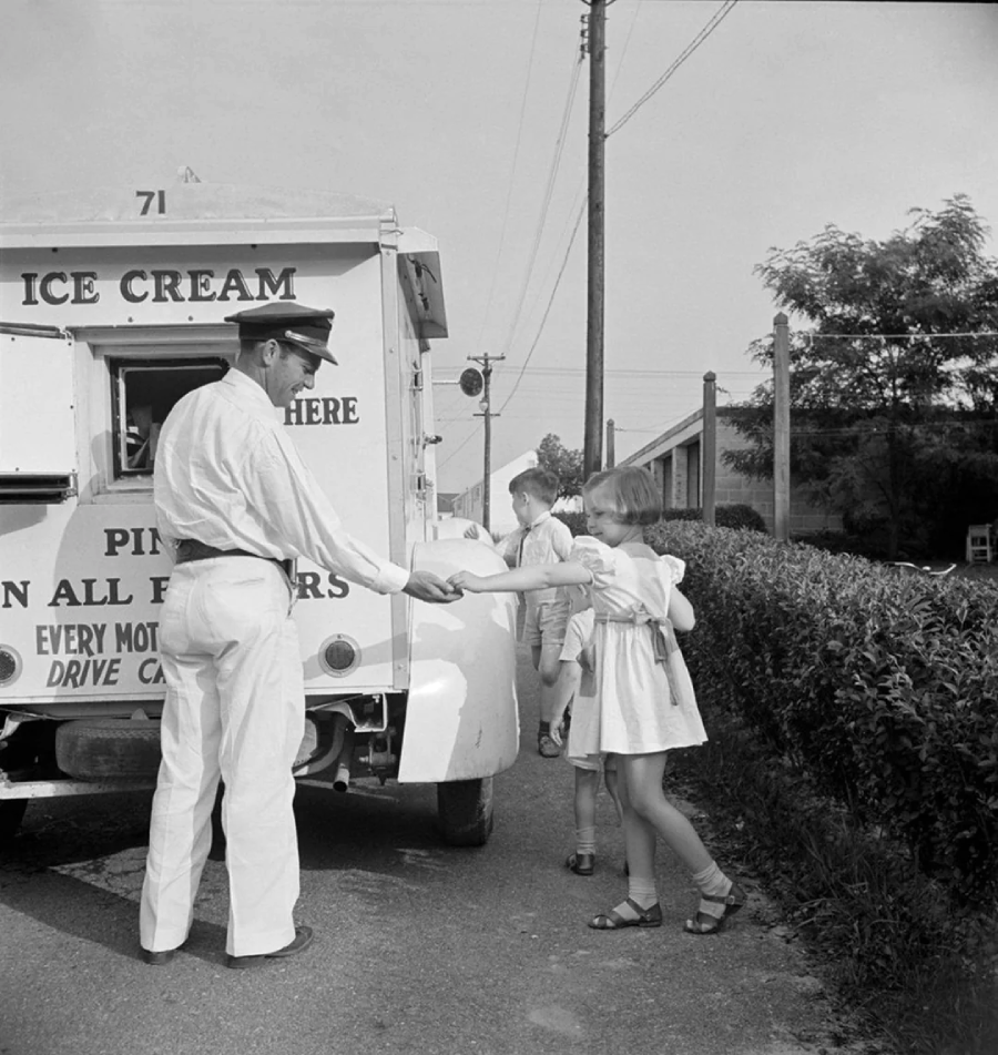 A man in a uniform serves ice cream from a truck to two children, a girl and a boy, who stand on a sidewalk next to the vehicle on a sunny day.