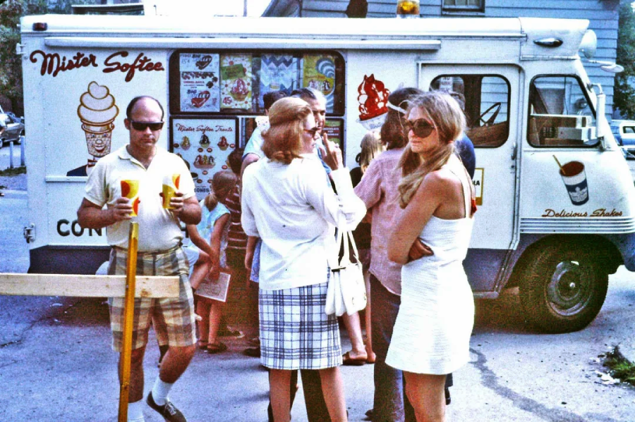 A group of people stands in front of a Mister Softee ice cream truck on a sunny day. Some hold ice cream or drinks, and the truck displays colorful menu signs. Everyone is casually dressed in summer clothing.