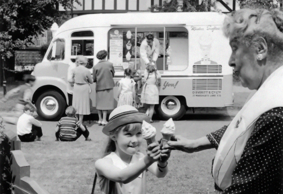 A young girl smiles while receiving an ice cream cone from an elderly woman in front of an ice cream truck, with several people and children gathered around the truck in the background.