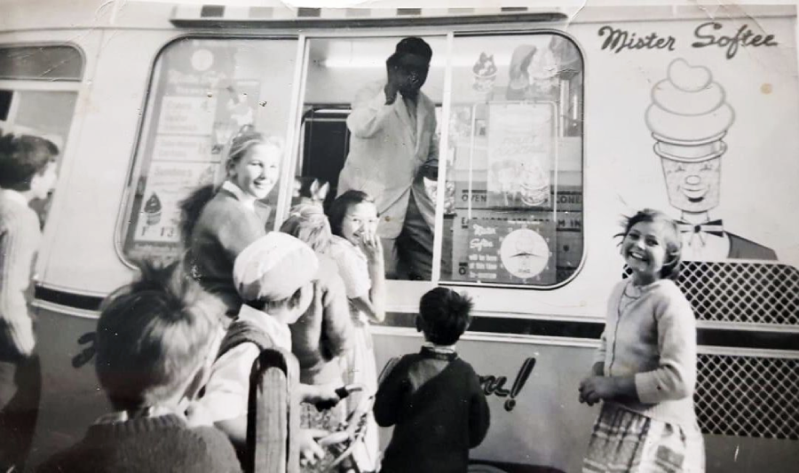 A black-and-white photo shows a group of happy children gathered around an ice cream truck labeled "Mister Softee," with a man inside the truck serving them. The kids are smiling and laughing.
