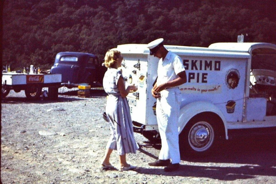 A woman in a striped dress stands beside a man in a white uniform and cap, next to a vintage Eskimo Pie ice cream truck parked on a gravel lot with trees and an old truck in the background.
