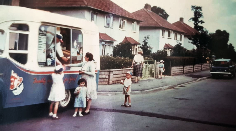 Children and adults gather around an ice cream van parked on a residential street lined with houses, as some receive treats on a sunny day. A classic car is parked further down the road.