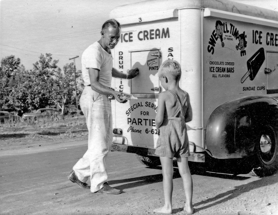 A man serves ice cream to a young boy standing beside a vintage ice cream truck on a sunny day. The truck displays signs for various ice cream treats and has a phone number for party services.