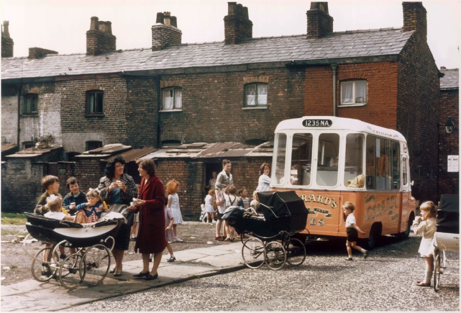 A vintage ice cream van is parked on a street in front of old brick houses. Several women and children, some with prams, gather around the van, enjoying treats and chatting on a sunny day.