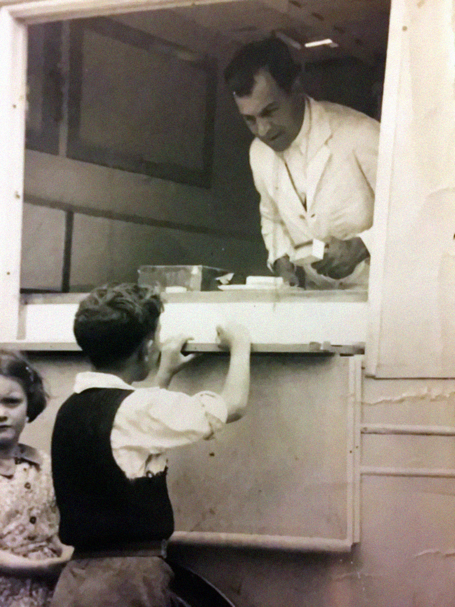 A black-and-white photo shows a man in a white coat serving two children from a window counter, possibly an ice cream or food truck. The boy stands at the counter while a girl stands nearby looking at the camera.