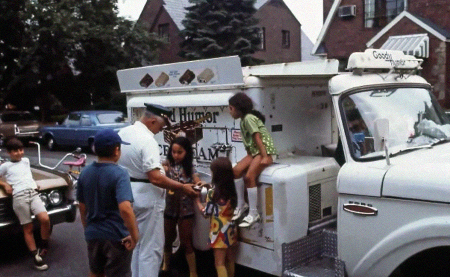 A man in a white uniform serves ice cream to children gathered around a classic Good Humor truck parked on a suburban street lined with houses and cars.