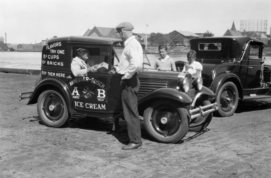 A vintage ice cream truck with “Anheuser-Busch Ice Cream” on the door is parked on a cobblestone street. The driver serves three men and boys, with another classic car parked nearby and buildings in the background.