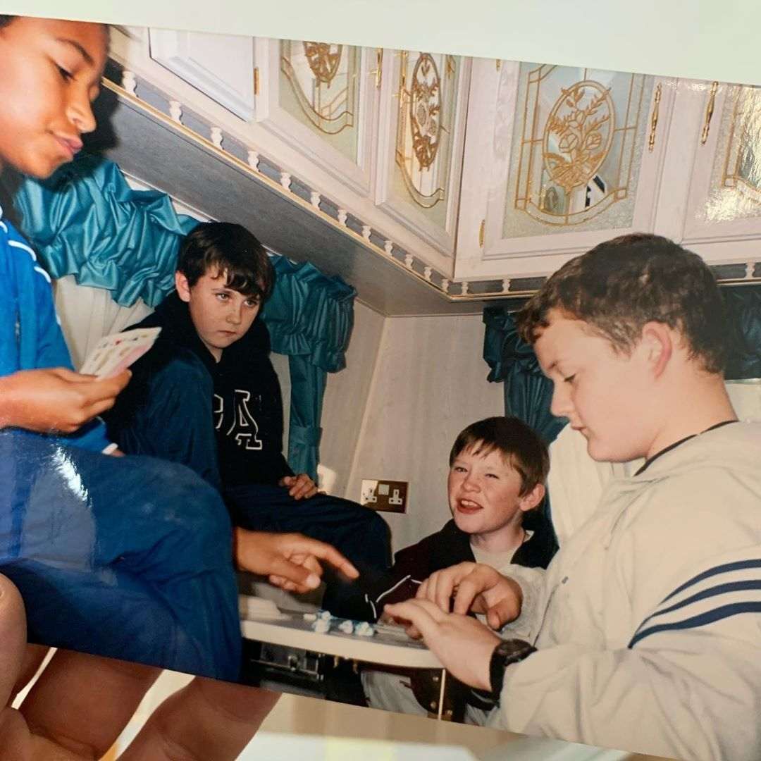 Four boys sit around a small table inside a RV or caravan. Two are holding playing cards, while another is reaching toward game pieces or a board on the table. They all appear focused and engaged.