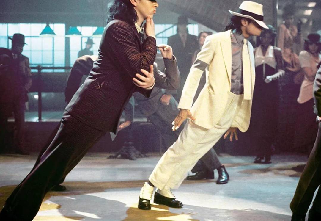 A group of dancers in suits perform a dramatic forward lean move on a dance floor, with one person in a white suit and fedora leading in the center. The scene appears energetic and stylized.