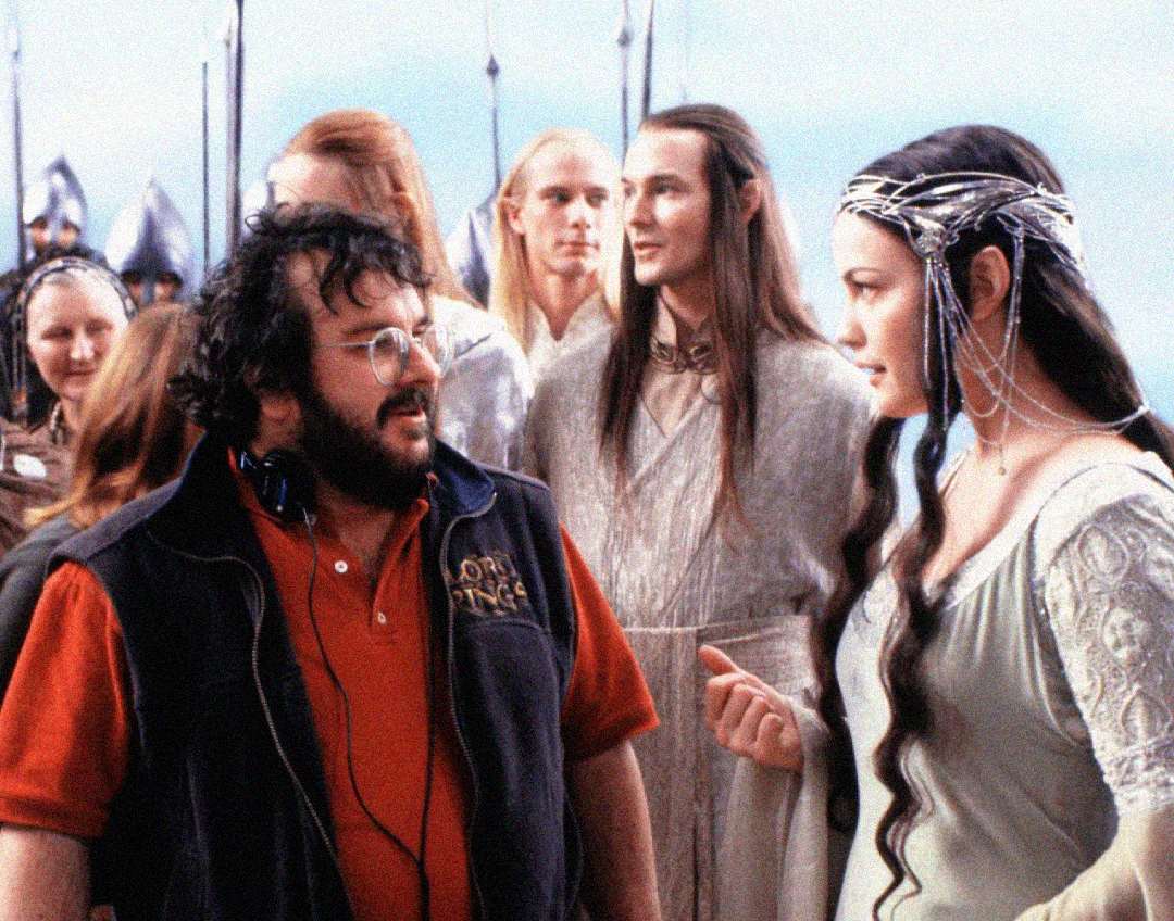 A man in a “Lord of the Rings” vest speaks with a woman in an elaborate medieval costume and headpiece, surrounded by other people in fantasy attire on a film set.