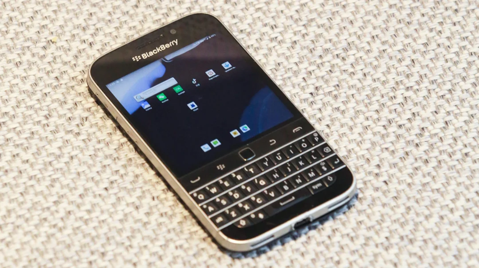 A BlackBerry smartphone with a physical QWERTY keyboard lies on a textured, light-colored fabric surface. The screen displays app icons and a faint reflection.