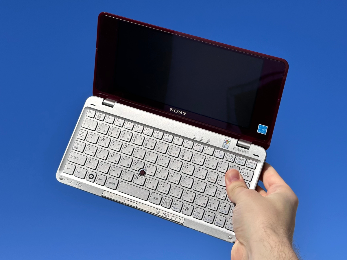 A hand holds a small, open Sony VAIO laptop against a clear blue background. The laptop has a silver keyboard and a dark screen.