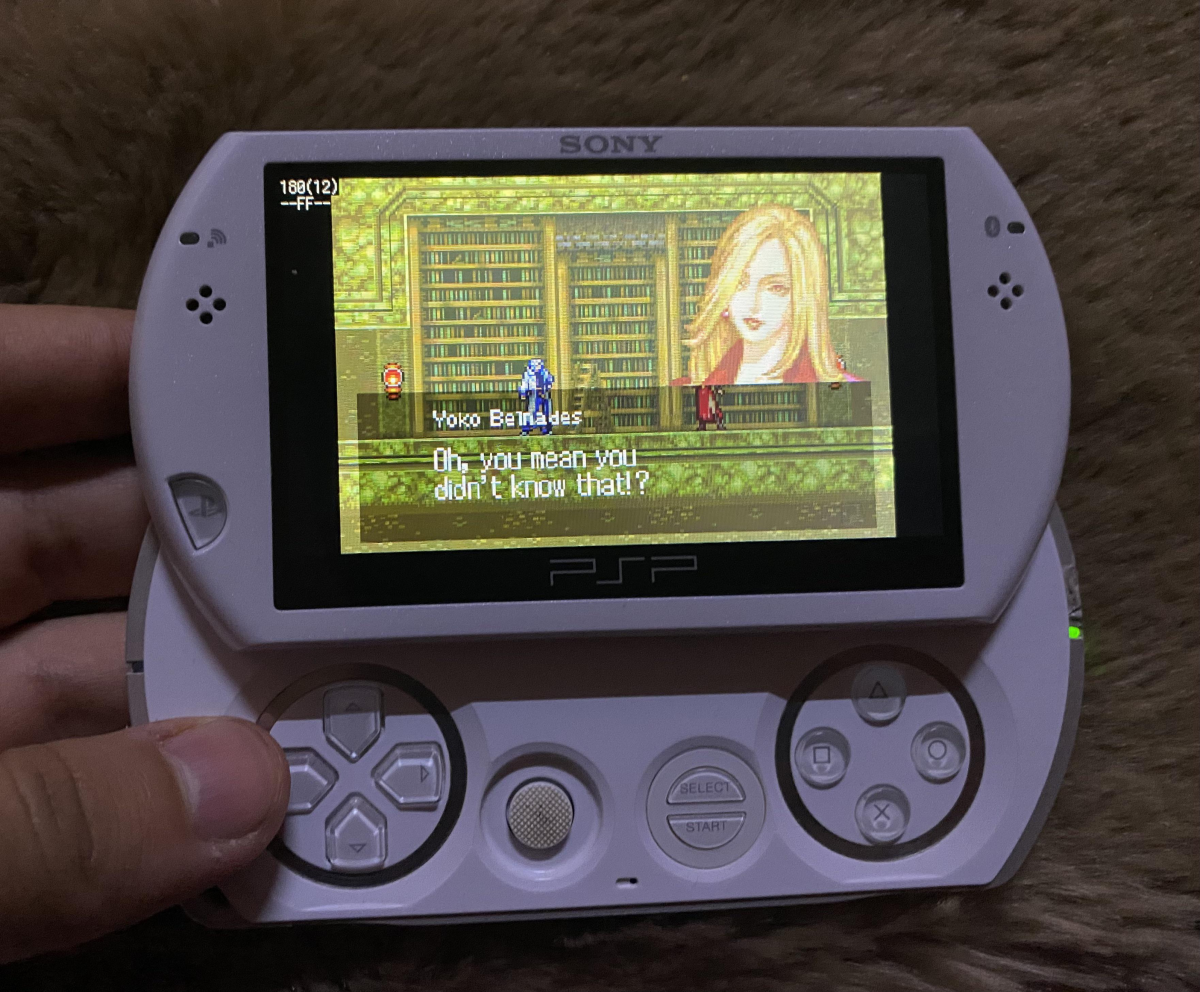 A person holds a white Sony PSP Go displaying a pixel art game scene with a dialogue box. The game shows a blonde female character and a player character standing in front of bookshelves.