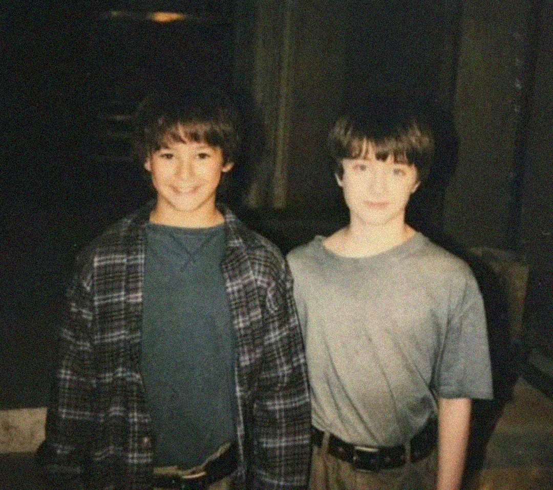 Two young boys with brown hair stand side by side, smiling for the camera. One wears a plaid shirt over a t-shirt, and the other wears a plain t-shirt. The photo has a slightly faded, vintage appearance.