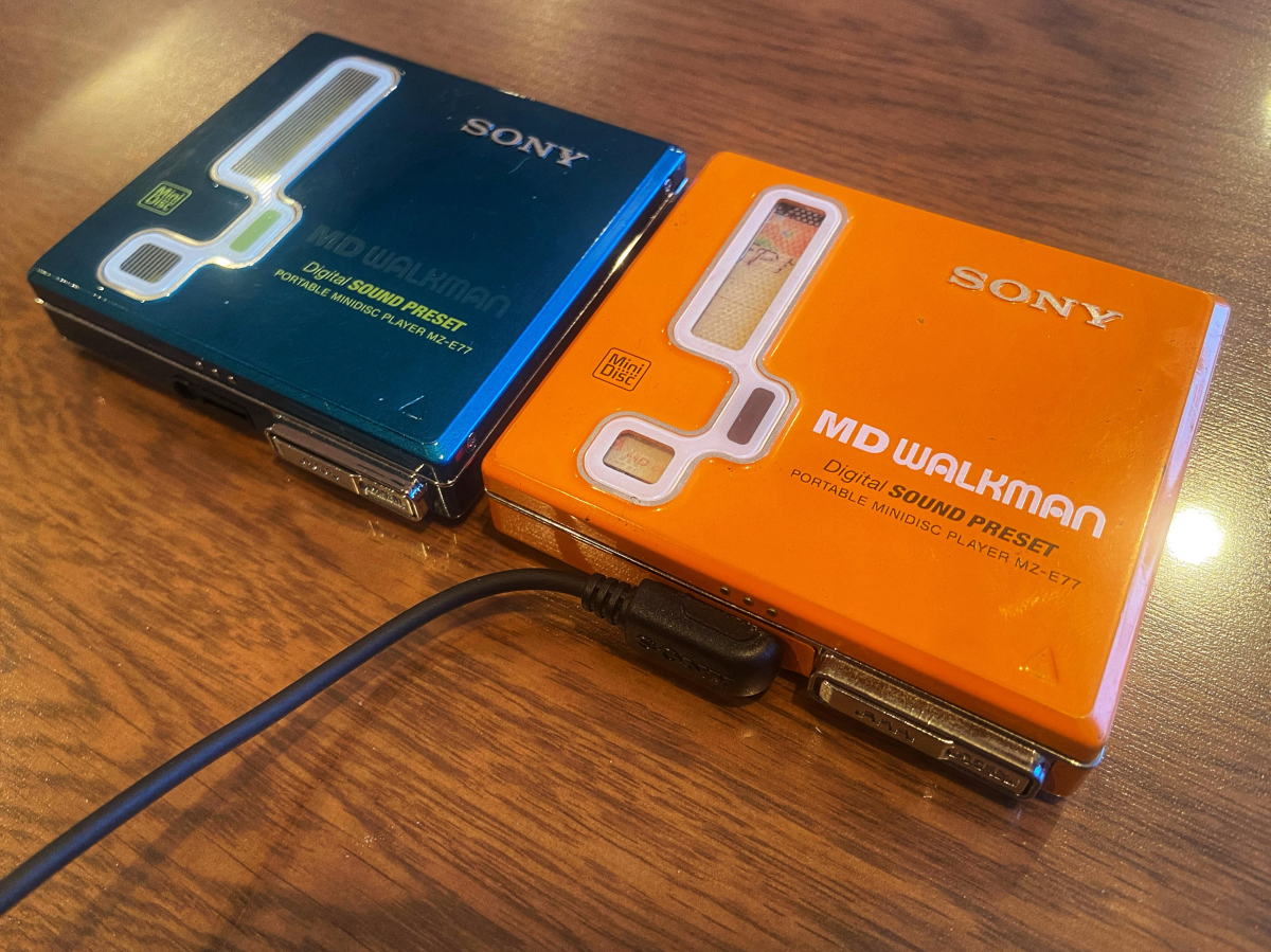 Two Sony MD Walkman MZ-E77 portable minidisc players, one teal and one orange, lie on a wooden surface. A black cable is plugged into the orange player.