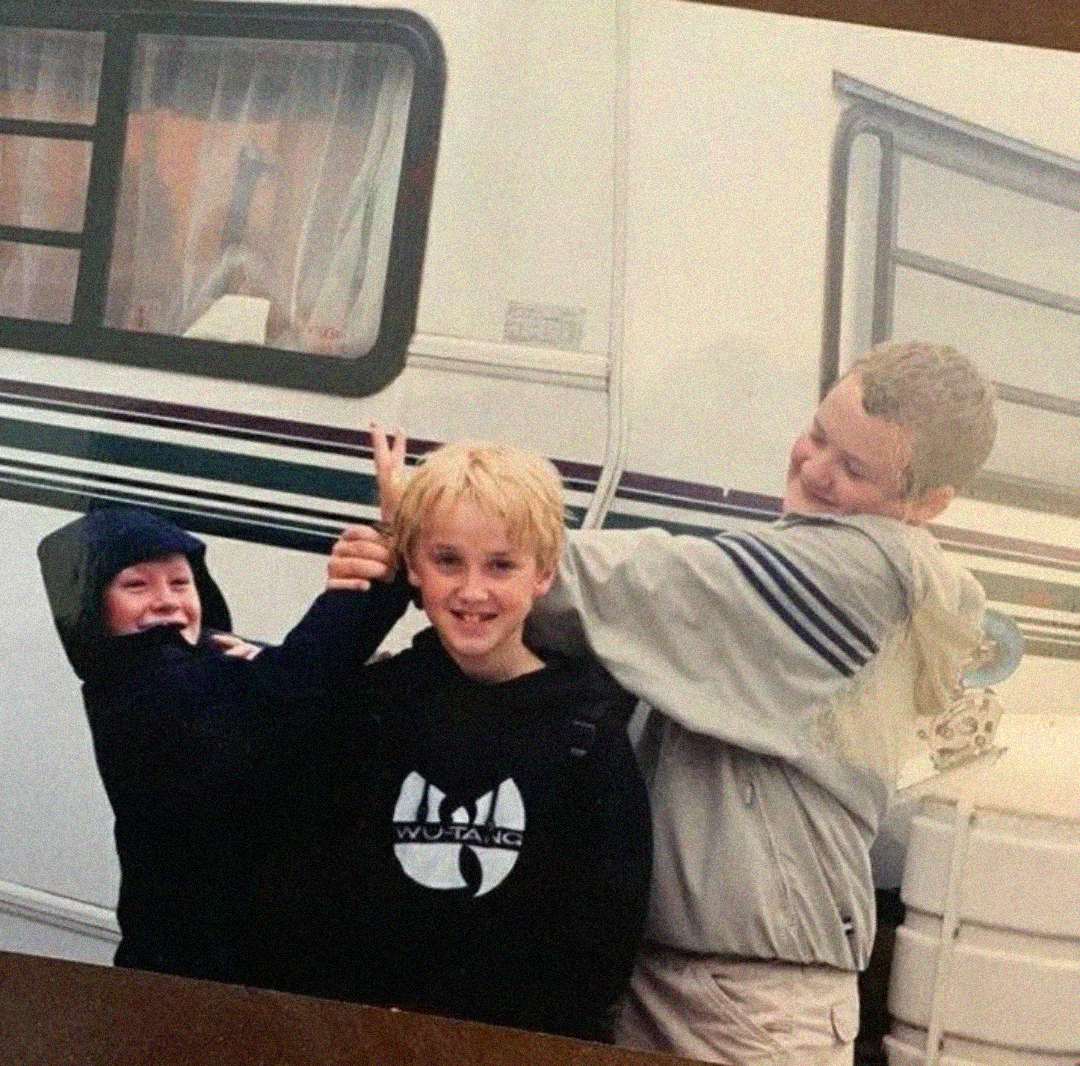 Three children are playfully posing in front of a camper van. One boy in a Wu-Tang Clan hoodie smiles at the camera, while another boy hugs him from the side, and a third child makes bunny ears behind his head.