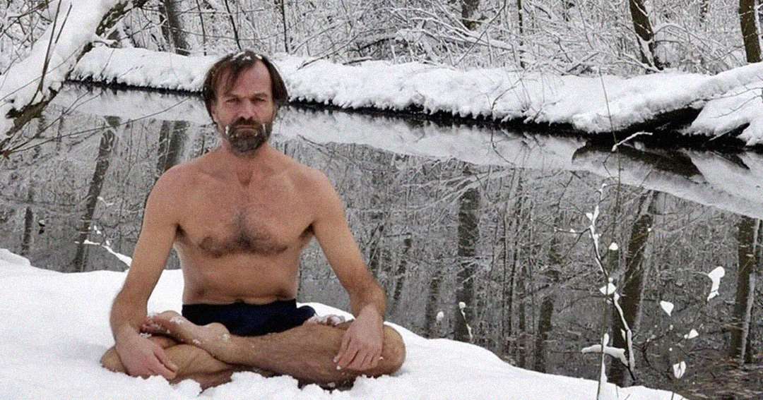 A man sits cross-legged in the snow beside a river, wearing only shorts. Snow-covered trees and a snowy landscape surround him, and he appears calm despite the cold.