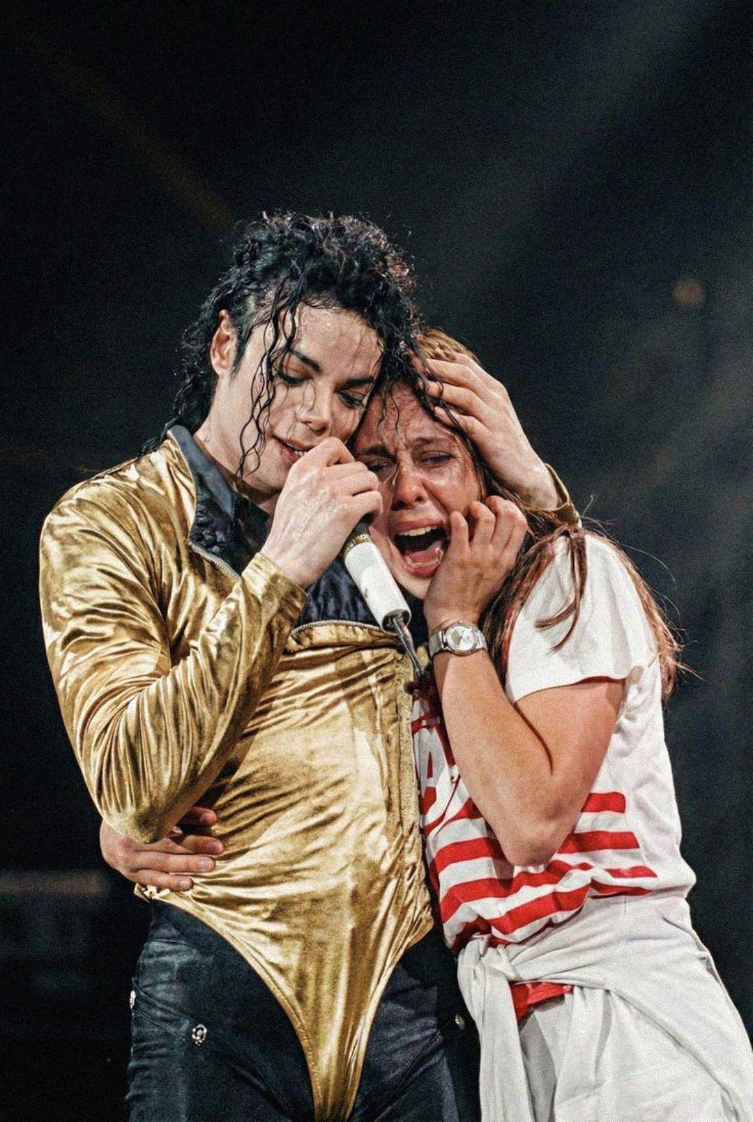 A singer in a gold outfit gently comforts a crying fan wearing a white and red striped shirt while performing on stage with a microphone.