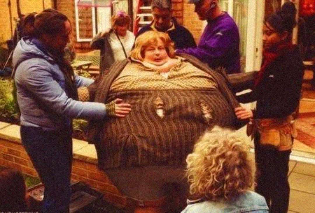 A person wearing an oversized, padded costume resembling an inflated suit is surrounded by several people adjusting the outfit outdoors on a film set.