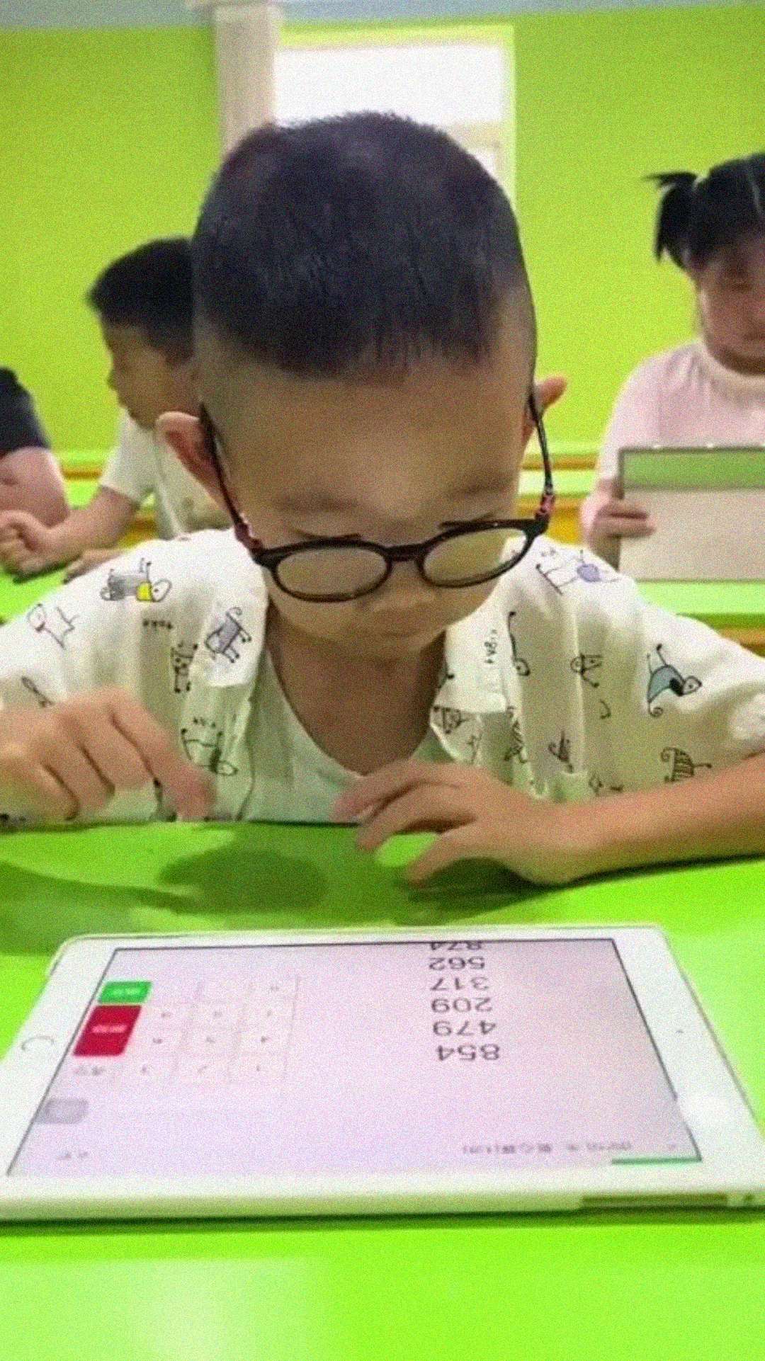 A young boy with glasses is focused on a tablet displaying math problems, seated at a bright green desk in a classroom with other children using tablets in the background.