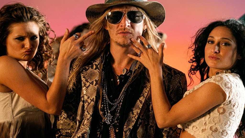A man in sunglasses, a snakeskin-patterned jacket, and a cowboy hat stands confidently between two women, who are posing with their hands near his face against a colorful background.