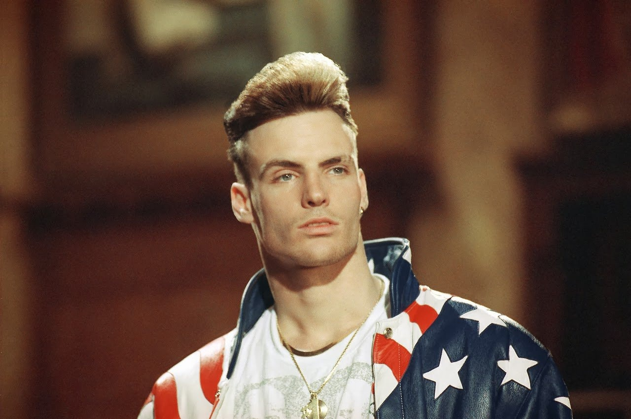A man with a tall, styled blond flattop haircut wears a jacket featuring the American flag pattern, looking off to the side in a dimly lit, indoor setting.