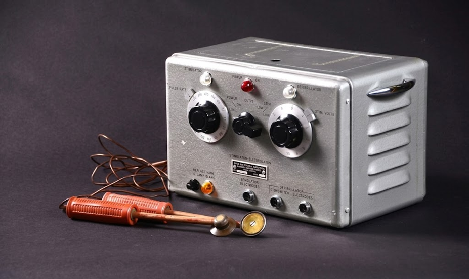 A vintage medical device with two analog dials, switches, indicator lights, attached cords, and handheld metal electrodes, set against a dark background.
