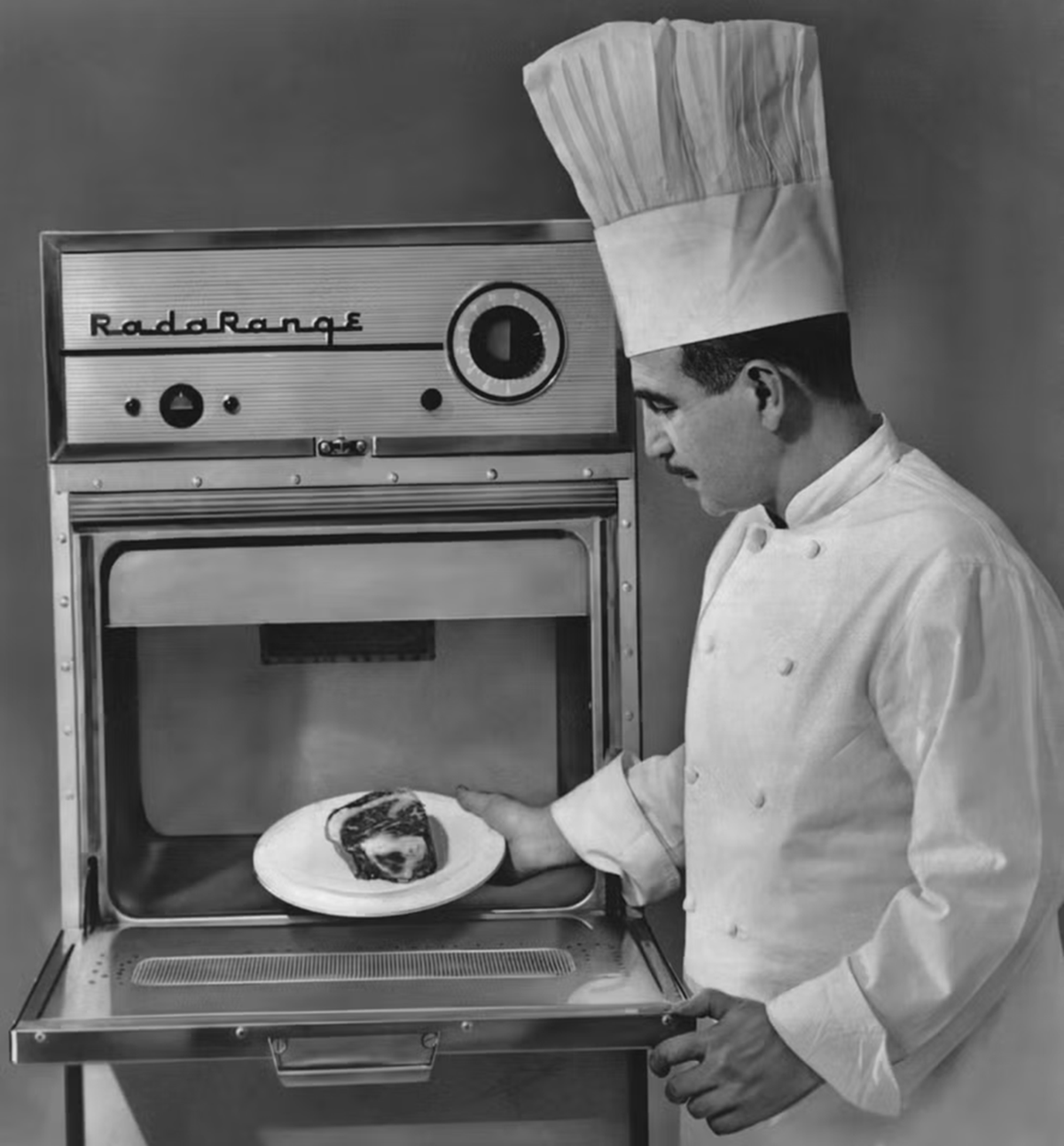 A chef in a white uniform and tall hat places a plate with a slice of meat or pie into a vintage Radarange microwave oven, with the oven door open and controls visible on the front panel.