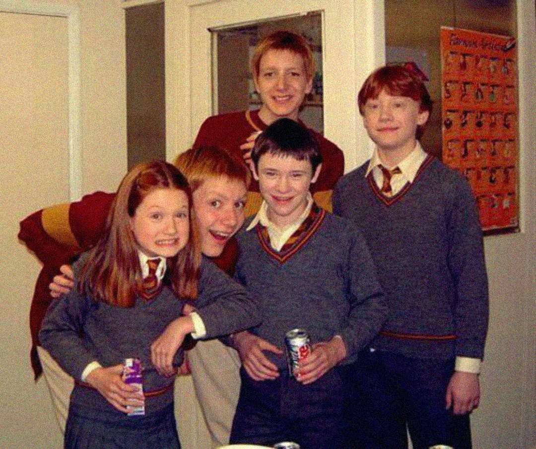 Five children in school uniforms smile at the camera, holding drinks. They are indoors, in what looks like a kitchen or break room with white walls and a calendar in the background.