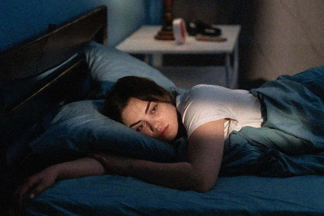 A person lies on a bed under blue sheets, resting their head on one arm and staring thoughtfully into the distance. The room is dimly lit, with a nightstand and objects visible in the background.