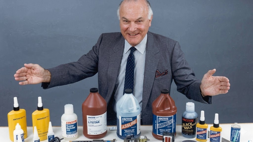 An older man in a suit smiles with arms open wide behind a table displaying various Loctite adhesive products, including large and small bottles and tubes, against a plain background.
