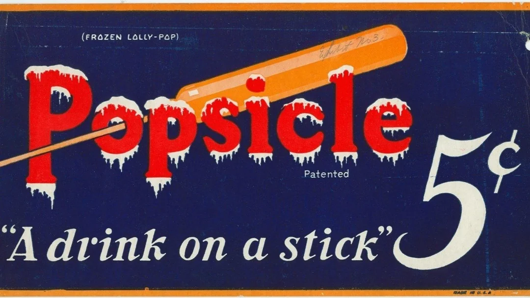 Vintage advertisement for Popsicle shows an orange popsicle on a stick over the large word "Popsicle," with the phrases "Frozen Lolly-Pop," "A drink on a stick," and "5¢" on a dark blue background.