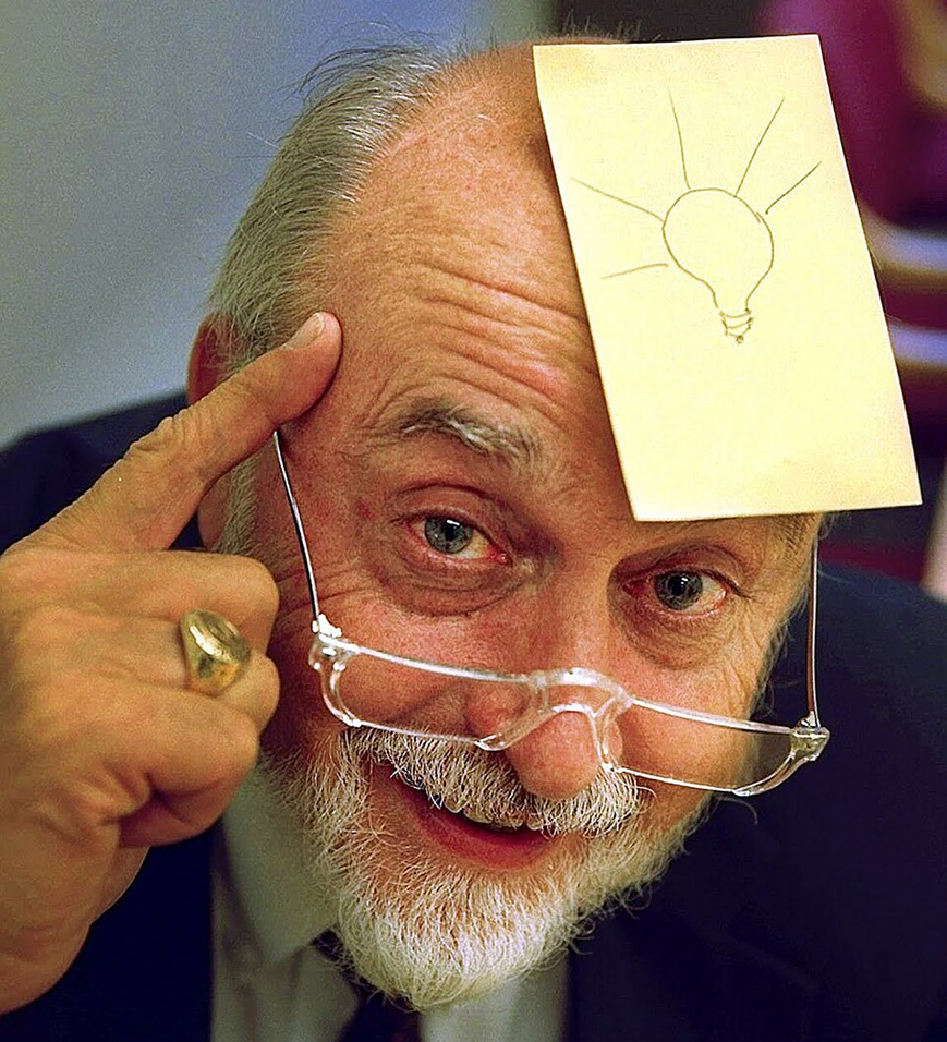 An older man with a white beard smiles, pointing to his temple. He has glasses and a sticky note with a drawing of a light bulb stuck to his forehead, suggesting he has an idea.