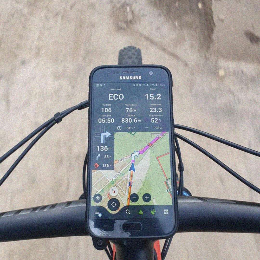 A smartphone is mounted on a bicycle handlebar, displaying a cycling app with speed, distance, route map, and navigation details. The bike is being ridden on a dirt path.
