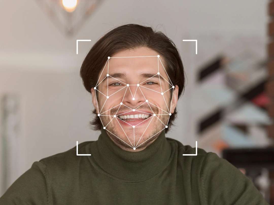 A smiling person with dark hair wearing a green turtleneck is facing the camera. White geometric lines and corner markers overlay their face, suggesting facial recognition technology in use.
