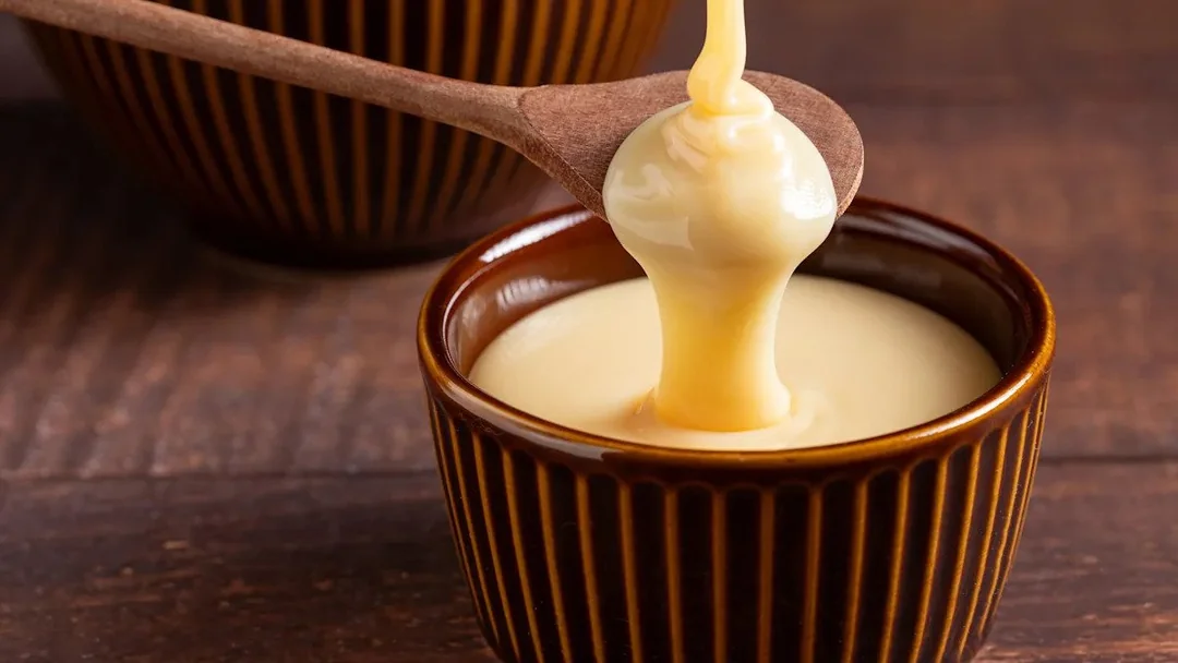 A spoon drizzling thick, creamy condensed milk into a small brown ceramic bowl with ridged sides, placed on a wooden surface.