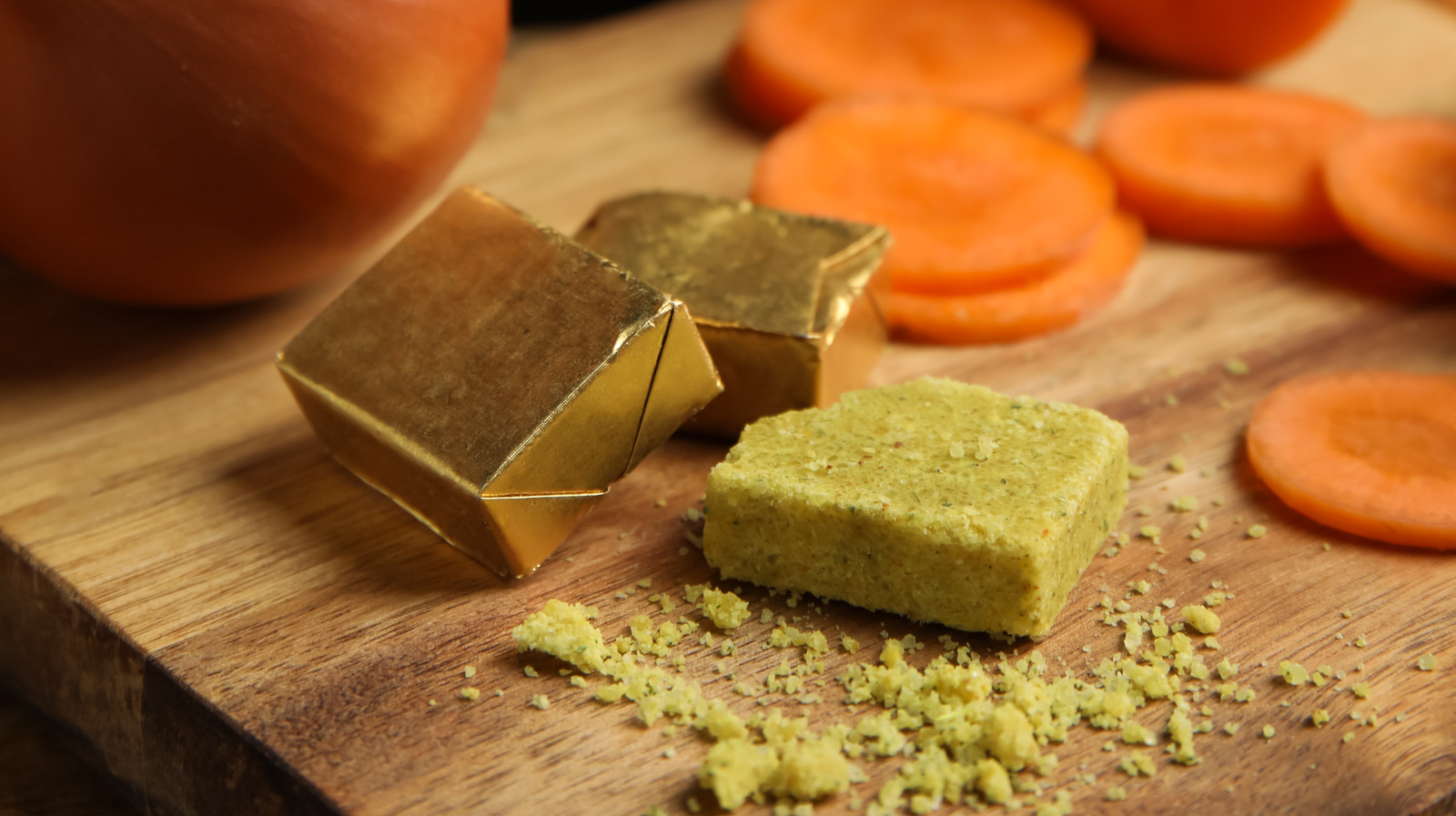 Vegetable bouillon cubes, one unwrapped and crumbling, sit on a wooden board next to sliced carrots and a partially visible orange vegetable, possibly a pumpkin.