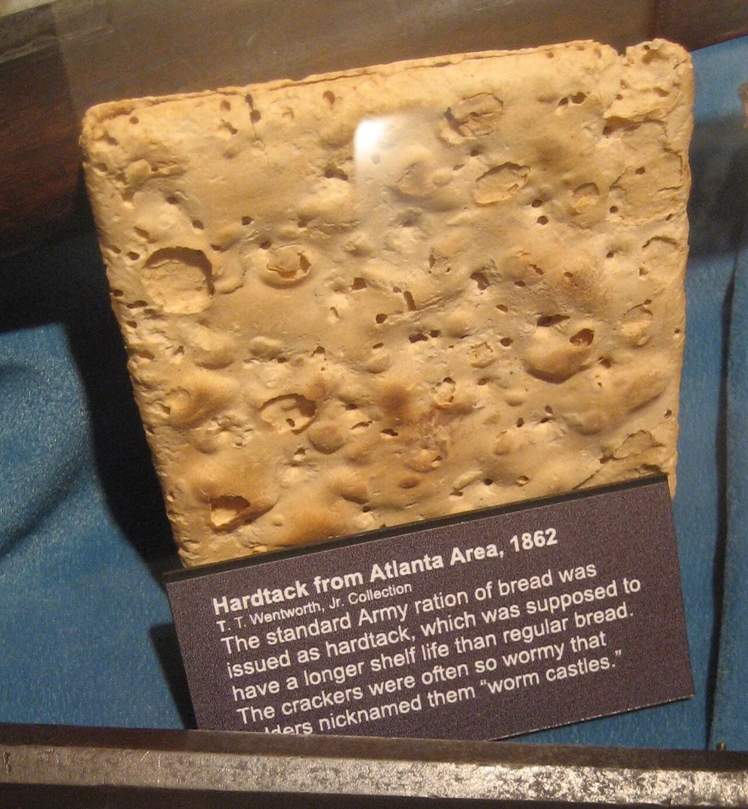 A square, rough, and pitted piece of hardtack bread sits on display with a descriptive label noting it is from the Atlanta area in 1862 and was a Civil War army ration known for its long shelf life and "wormy" appearance.