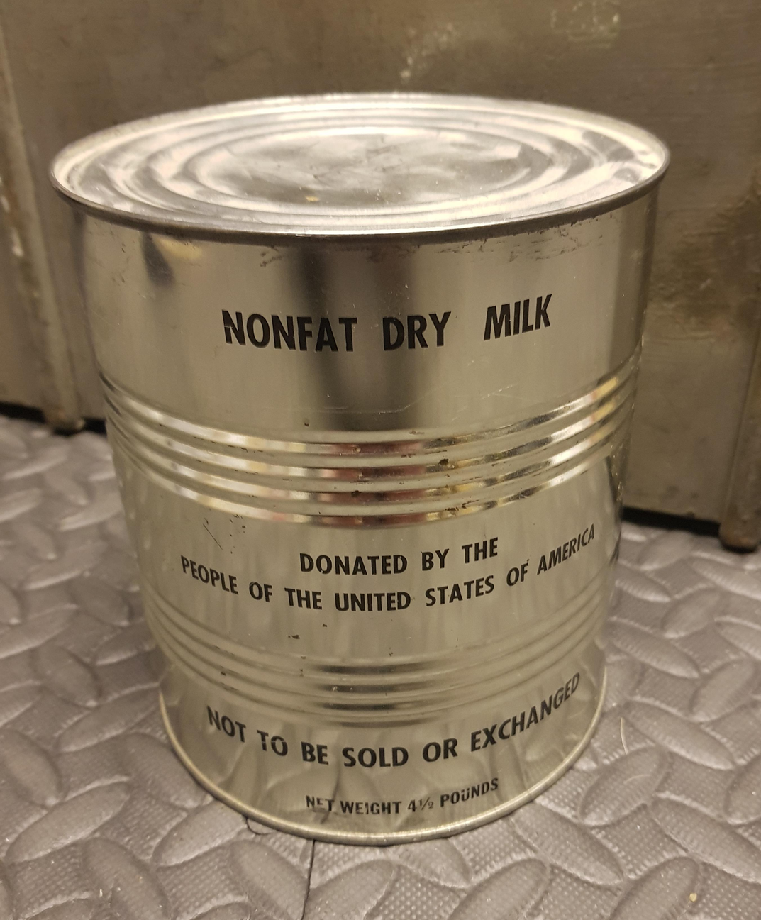 A large metal can labeled "Nonfat Dry Milk" with text stating it was donated by the people of the United States of America and is not to be sold or exchanged; net weight is 4½ pounds.