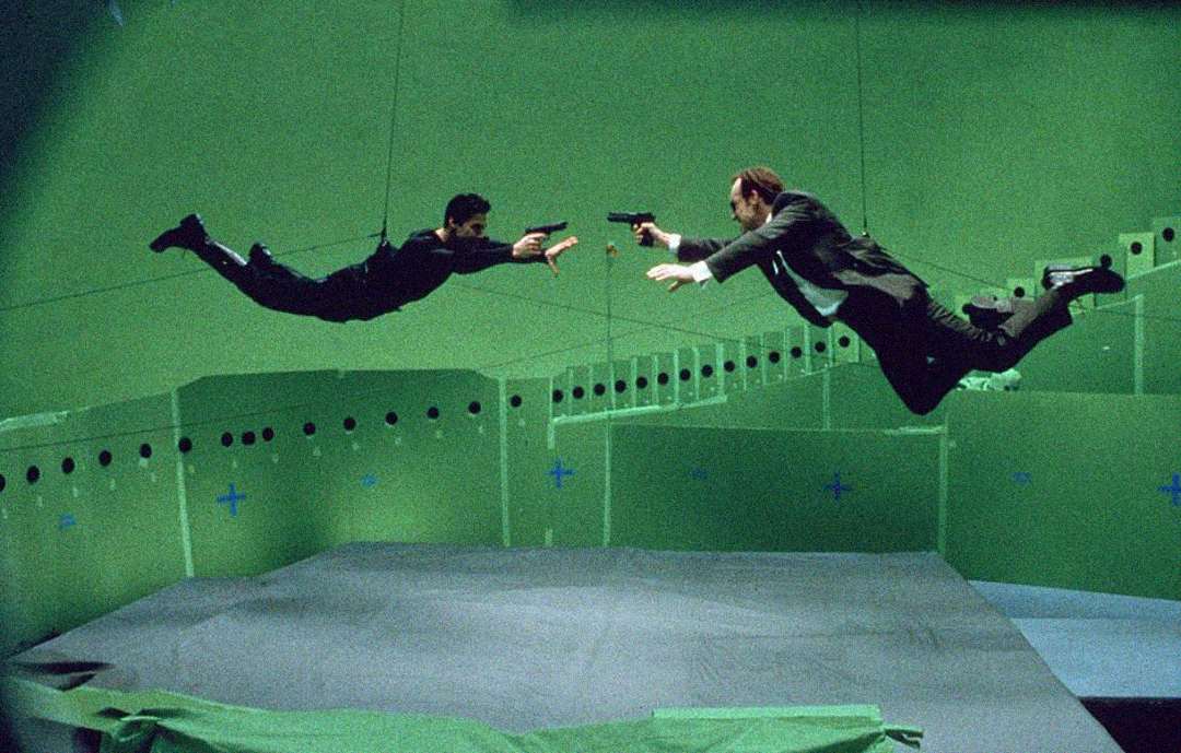Two actors in black clothing and a suit, suspended by wires, point guns at each other while floating in a green screen studio, simulating a dramatic action scene for a film.