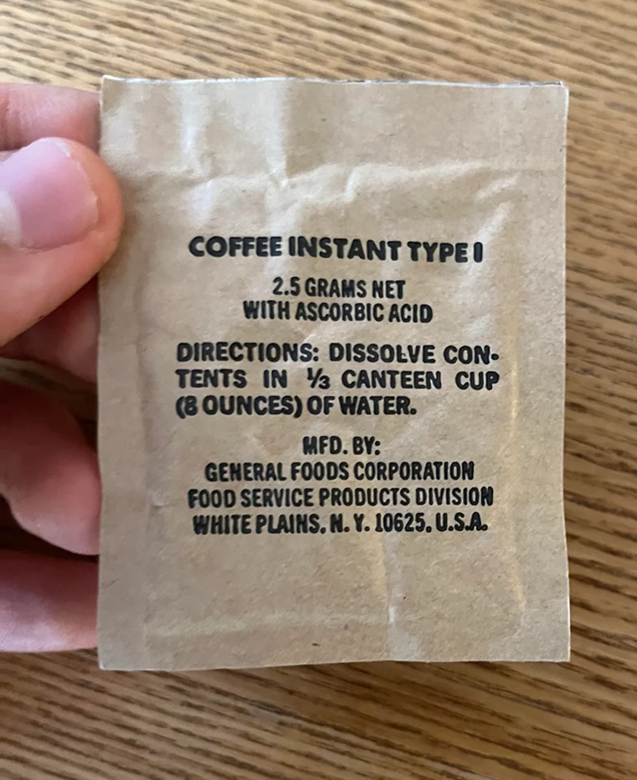 A hand holds a brown packet labeled "COFFEE INSTANT TYPE I" with instructions to dissolve the 2.5 grams of contents with ascorbic acid in 8 ounces of water. The packet is manufactured by General Foods Corporation.