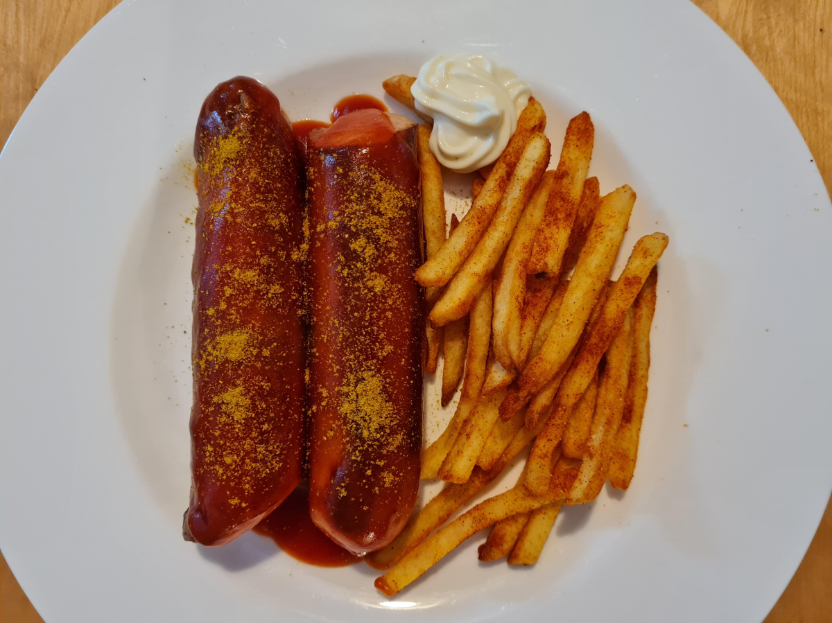 Two sausages topped with curry ketchup and curry powder, served with a side of seasoned French fries and a dollop of mayonnaise on a white plate.
