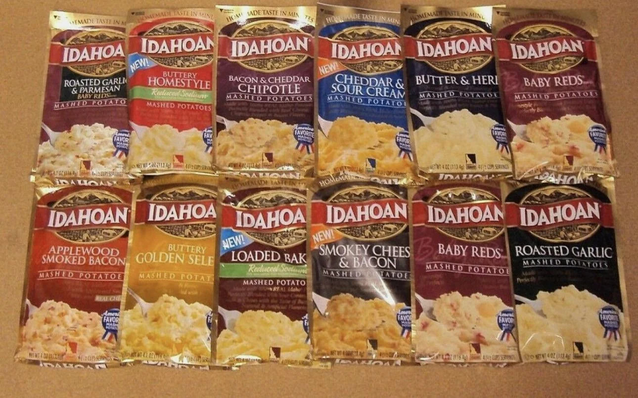 Twelve packets of Idahoan instant mashed potatoes in various flavors, including Roasted Garlic, Butter Homestyle, Cheddar & Sour Cream, Applewood Smoked Bacon, Loaded Baked, and Butter & Herb, arranged in two rows.