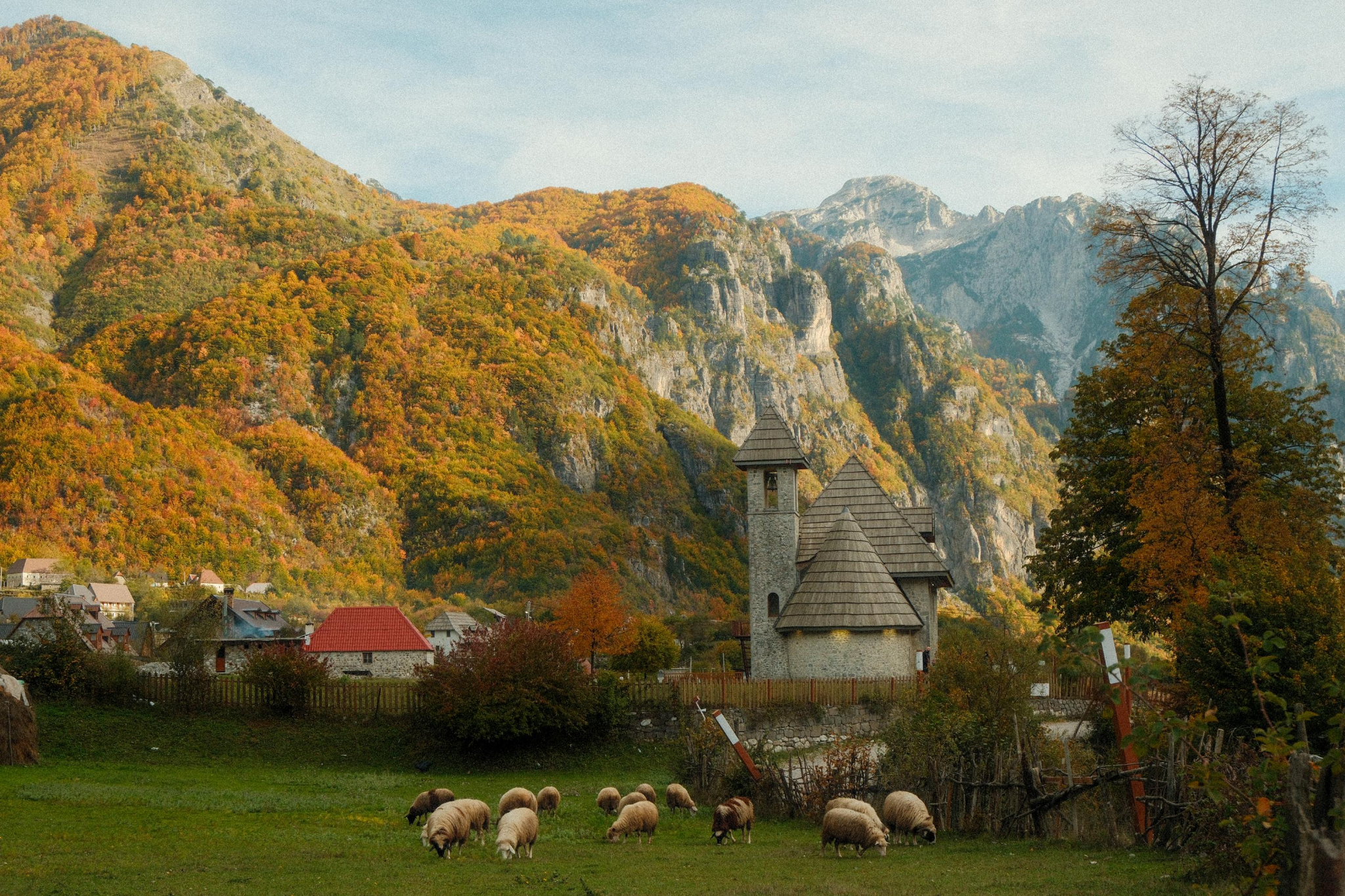 A small flock of sheep grazes on a grassy field in front of a stone church with towers, surrounded by colorful autumn trees and dramatic mountain cliffs in the background. Destinations