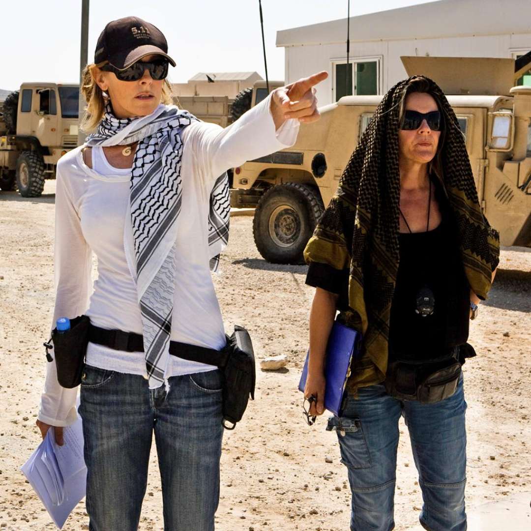 Two women in casual clothing and scarves stand outdoors near military vehicles; one points into the distance while holding papers, and the other holds a blue notebook and stands beside her. Both wear sunglasses and look focused.